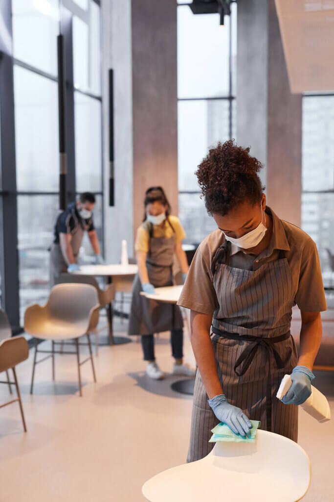 A young woman in a cafe diligently cleaning tables with a spray bottle labeled 3% food grade hydrogen peroxide.