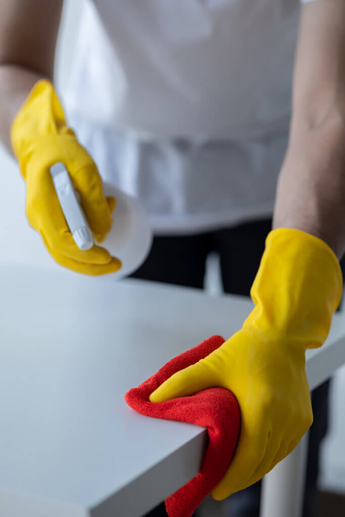 Individual dressed in white, wearing rubber gloves, holding a spray bottle of hydrogen peroxide, and cleaning a countertop.